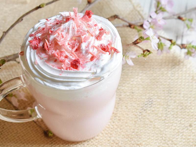 Mug of pink drink with whipped cream and red toppings on a textured surface with cherry blossom branches.