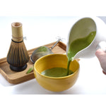 Pour matcha from a white-handled teapot into a yellow matcha bowl. A tea strainer, a bamboo whisk