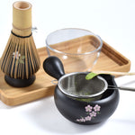 Tea-making set with black teapot, glass cup, and bamboo whisk on a white background