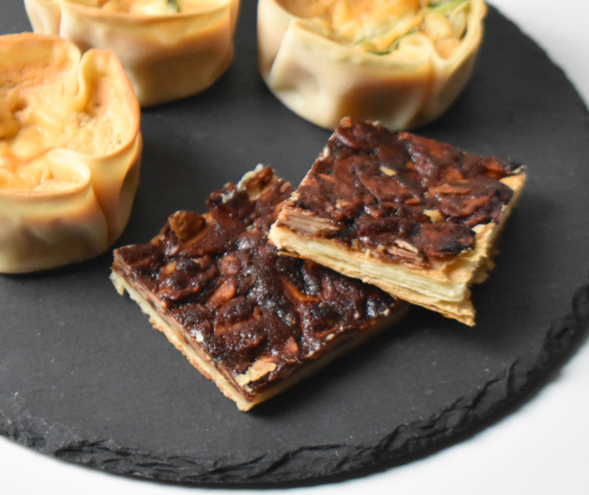 Two pecan pie bars on a black slate board with other pastries in the background.