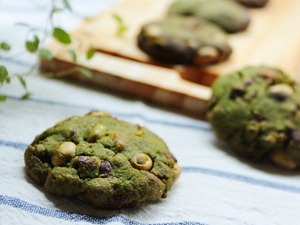 A close-up of a green chocolate chip cookie with pistachio pieces in the center, with more cookies on a wooden board in the background on a blue-and-white striped cloth.