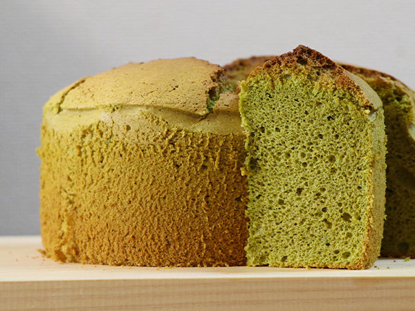 A sliced loaf of green cake, showing its moist crumb texture, resting on a light wooden board against a neutral gray background.