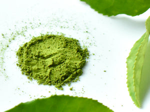 A small mound of bright green matcha powder sits on a white surface, surrounded by fresh green tea leaves.