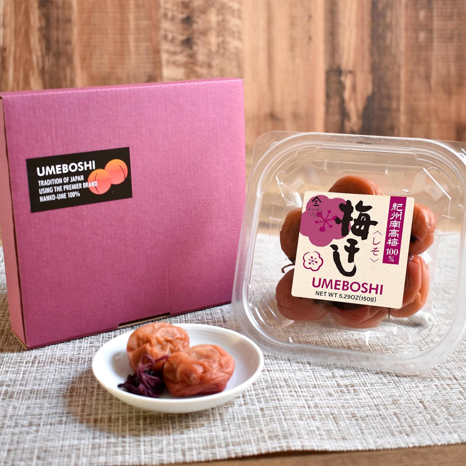 Umboshi packaging with pink box, clear container, and white plate on a wooden surface.
